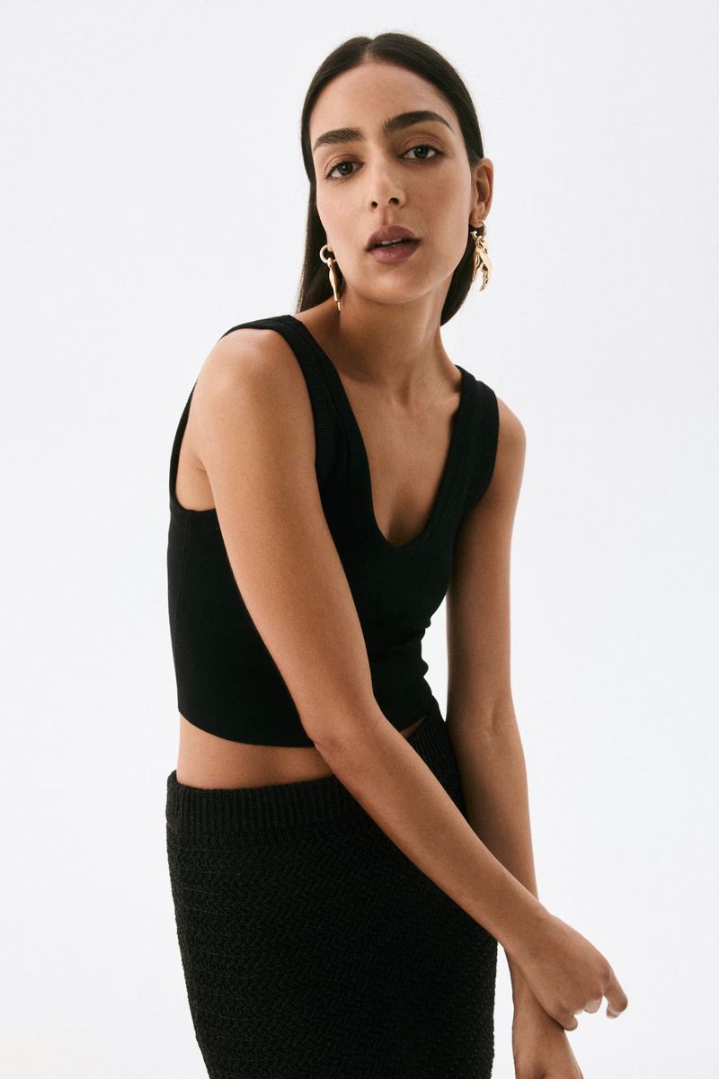 A woman with dark hair poses in a studio wearing a black top and textured skirt against a white backdrop, showcasing a minimalist aesthetic.