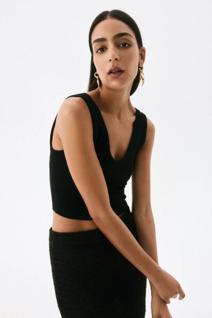 A woman with dark hair poses in a studio wearing a black top and textured skirt against a white backdrop, showcasing a minimalist aesthetic.