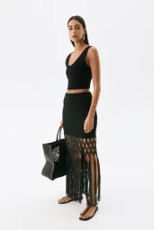 Model wearing a black crop top and fringed crochet skirt holding a large black tote bag against a white background.