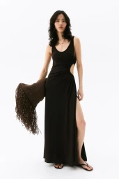 Model in a black maxi dress with side cutouts holding a fringed brown bag against a white background