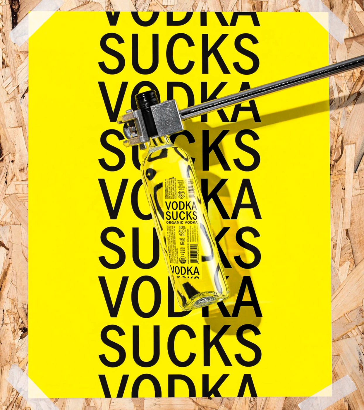 A yellow poster with repeated black text reading VODKA SUCKS attached to an OSB wood surface by white tape, with a clear glass bottle clamped by a metal tool diagonally across it.