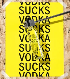 A yellow poster with repeated black text reading VODKA SUCKS attached to an OSB wood surface by white tape, with a clear glass bottle clamped by a metal tool diagonally across it.