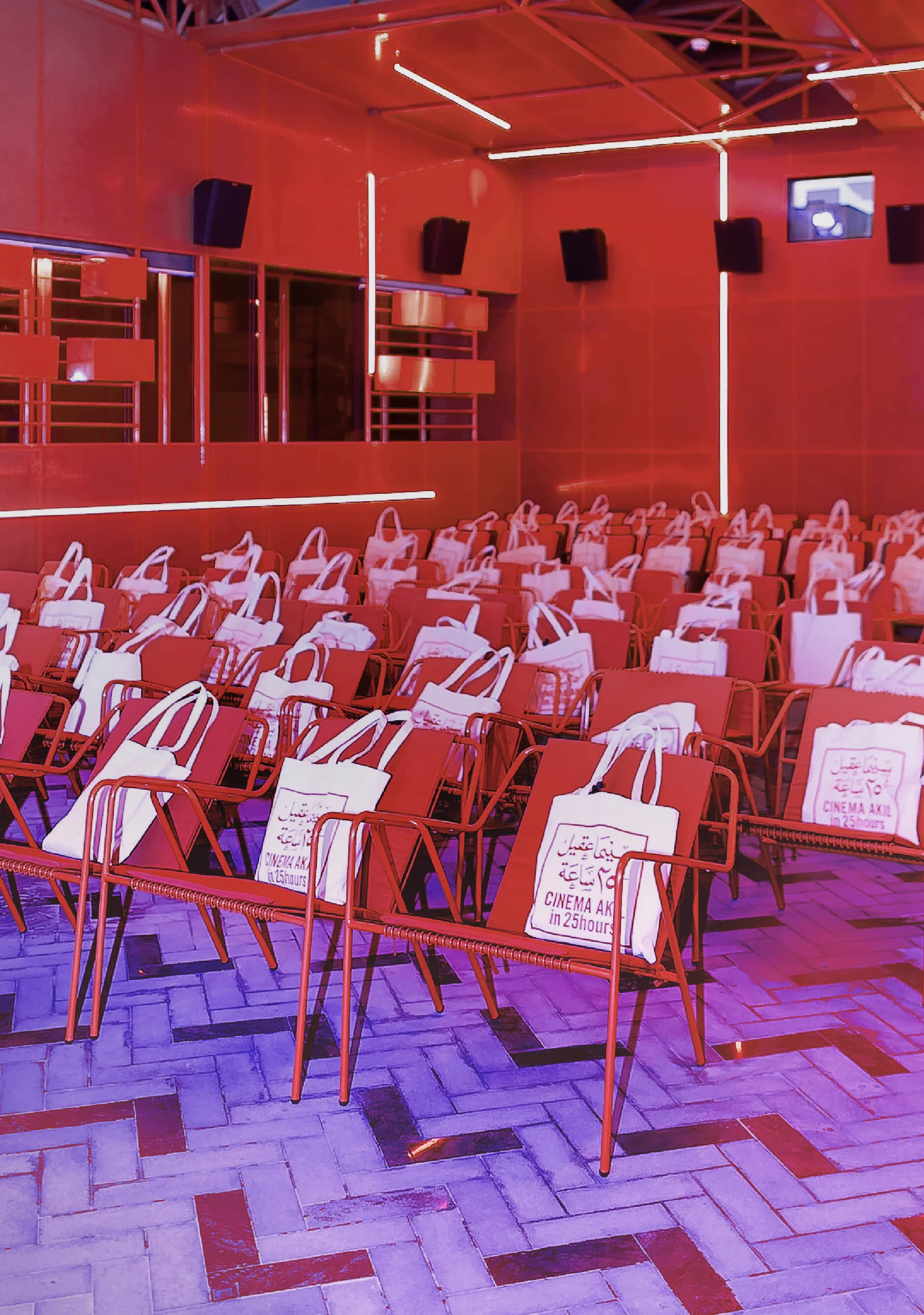 Indoor auditorium filled with rows of red metal chairs, each adorned with a white tote bag; overhead lighting illuminates the space.
