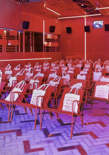 Indoor auditorium filled with rows of red metal chairs, each adorned with a white tote bag; overhead lighting illuminates the space.