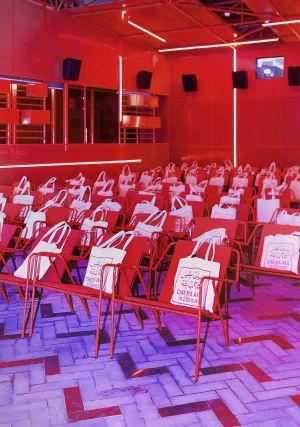 Rows of red metal cinema chairs inside a theatre bathed in dramatic red lighting, each seat holding a white tote bag.