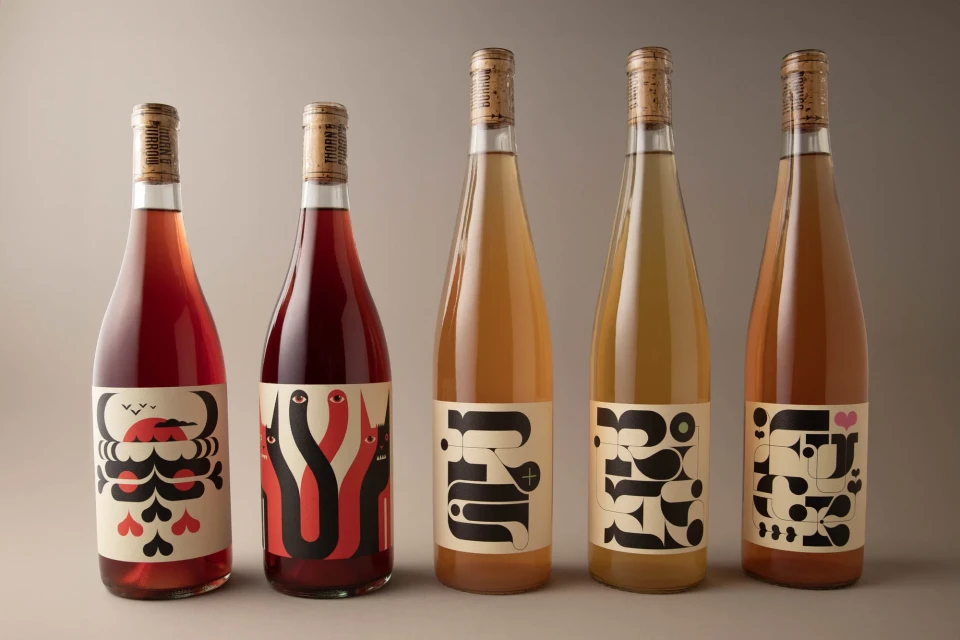 Five bottles of wine stand in a row, each with a unique, graphic label in red, black, and white against a soft, neutral background.