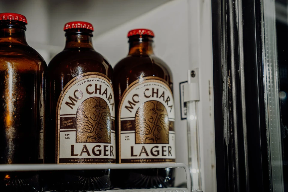 Three brown glass beer bottles with red caps on a white wire shelf inside a refrigerator with a black frame and door.