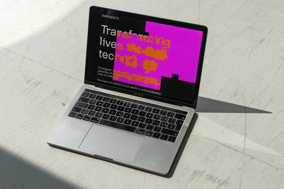 Laptop displaying website for EmergentX on a white surface with a shadow. Screen shows web copy about transforming lives through technology.