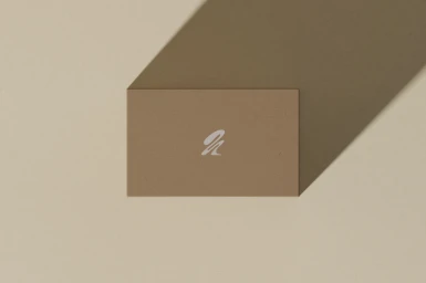 Tan business card featuring a minimalist white logo resting on a matching beige surface with a long shadow.