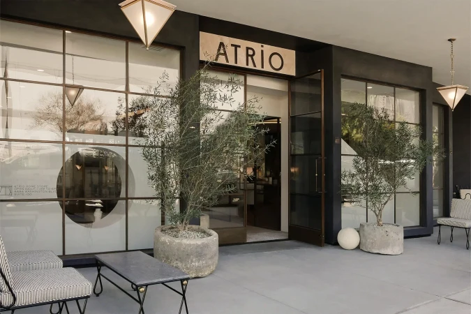 Exterior view of the Atrio store front with large windows, black trim, planters, chairs, and a table on a neutral tone walkway.