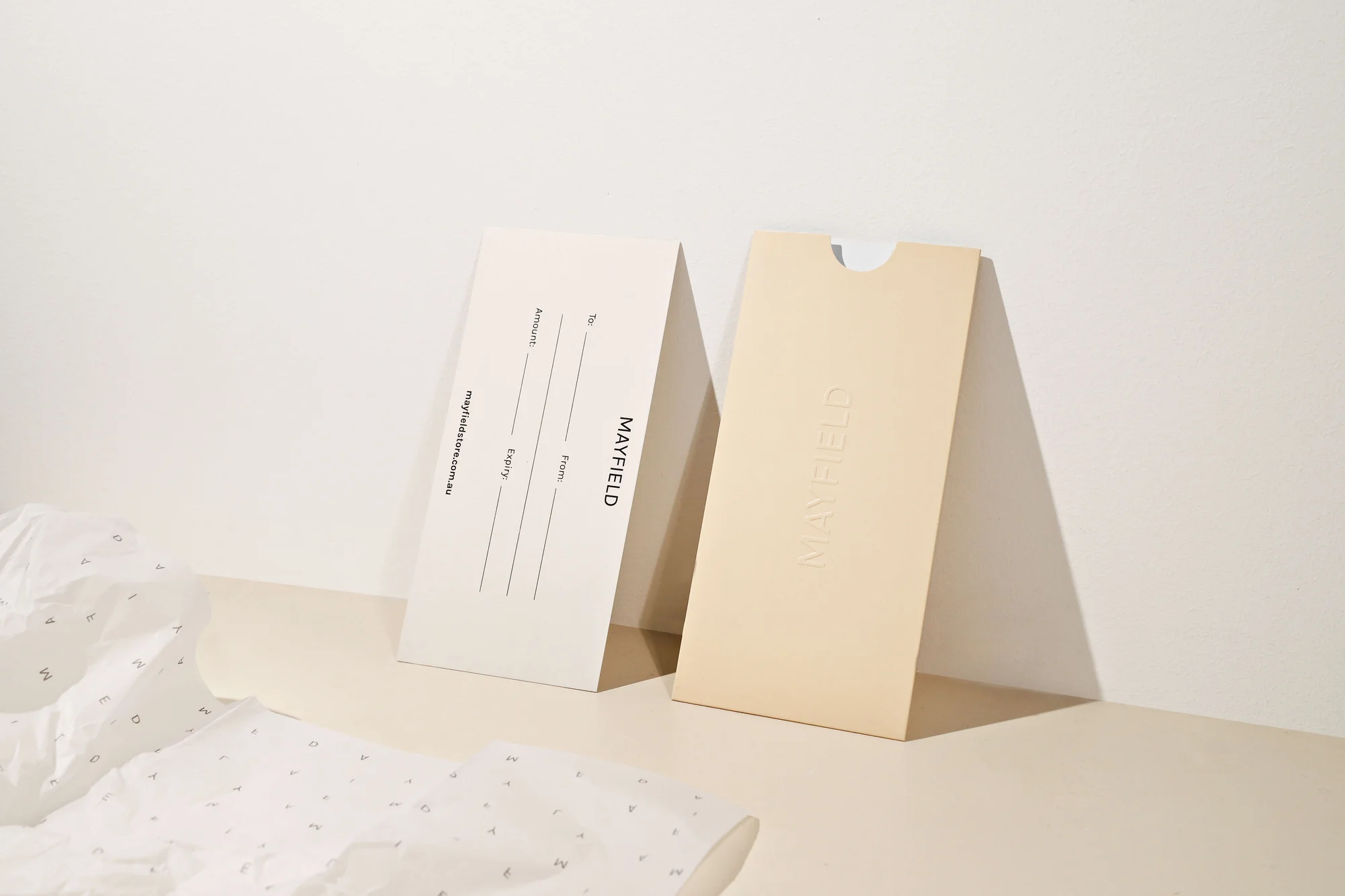 Two Mayfield gift card holders, one cream and one tan, rest on a light surface with patterned tissue paper and a neutral background.