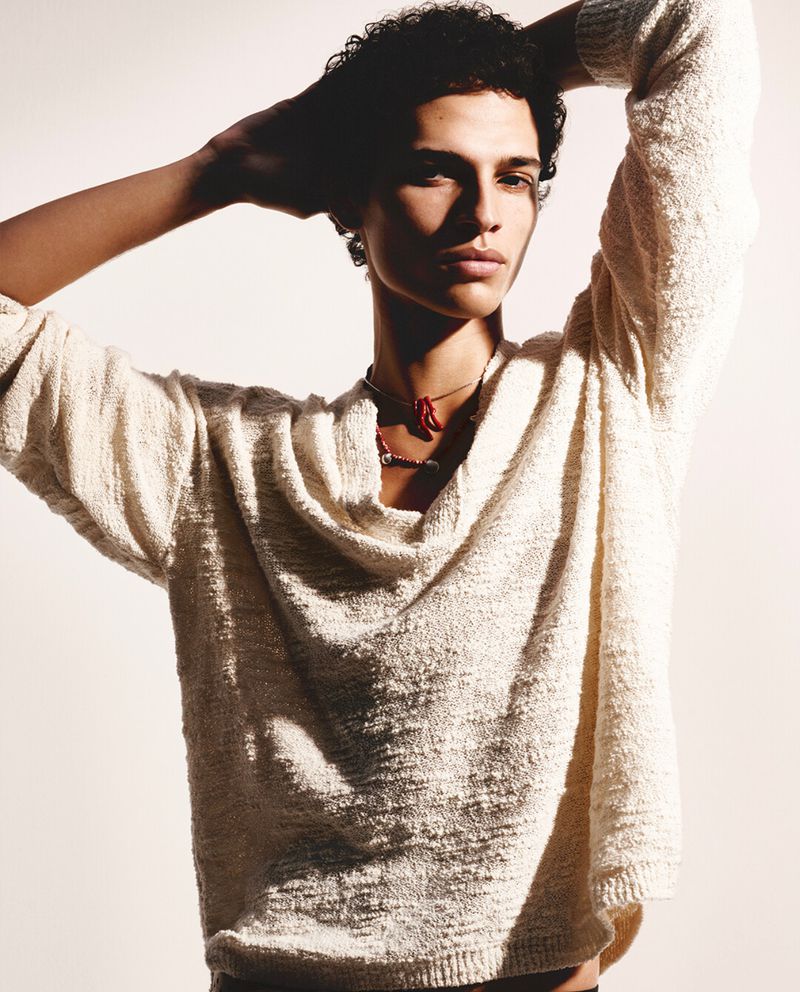 Portrait of a curly-haired male model wearing a cream-colored sweater, arms raised behind his head against a bright white background.