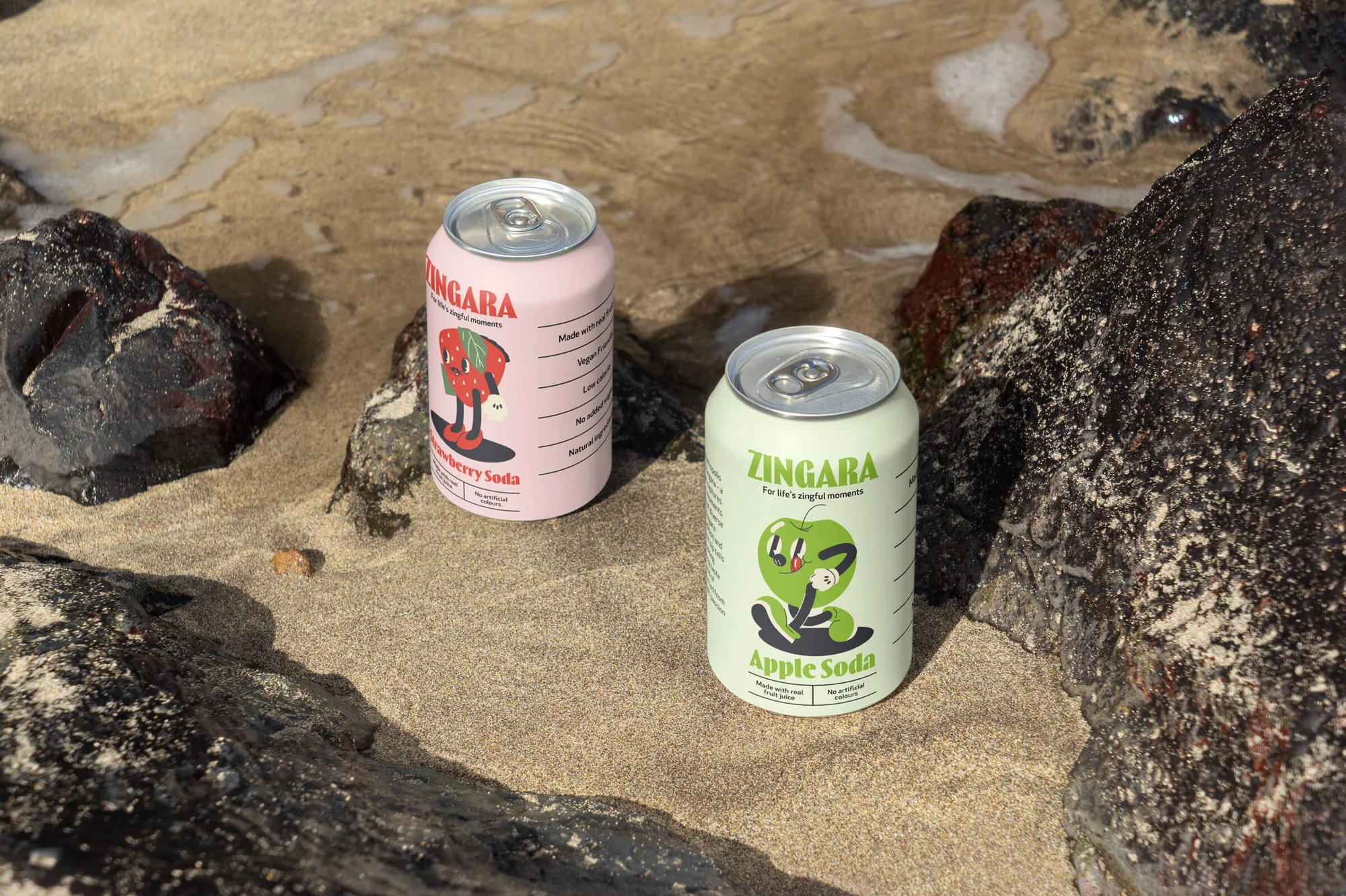 Two cans of Zingara soda, strawberry and apple flavor, sitting on a sandy beach next to dark rocks, with a water background.