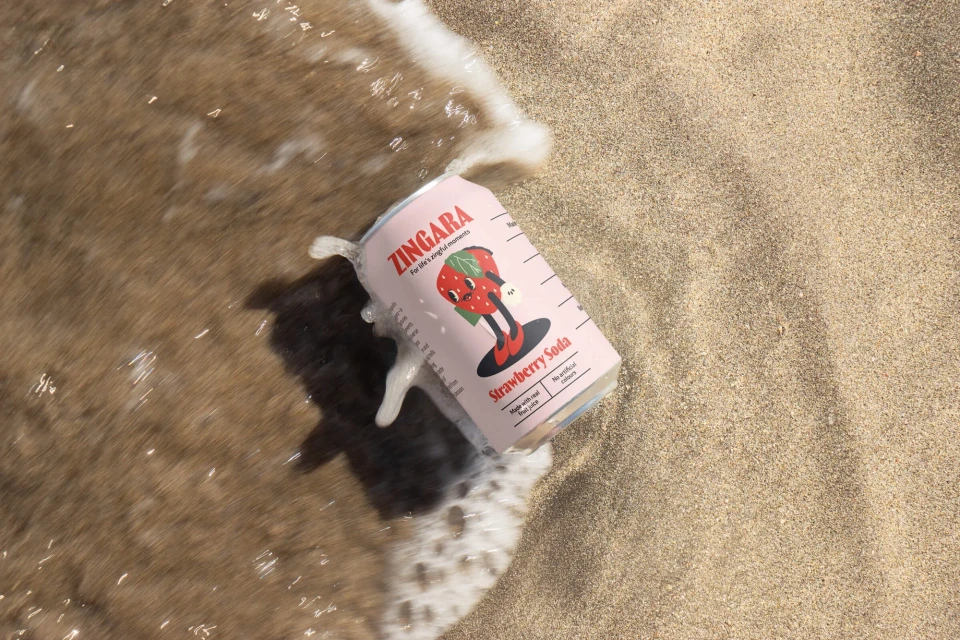 Strawberry soda can in wet sand by surf. Pink Zingara can with strawberry mascot in waves on beach. Food and beverage photography.