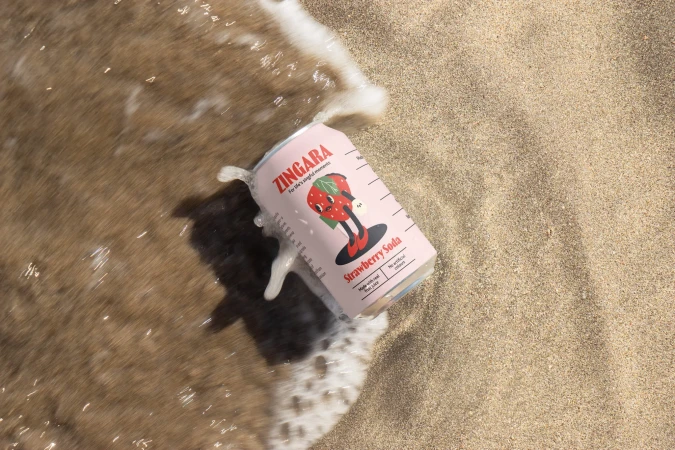 Strawberry soda can in wet sand by surf. Pink Zingara can with strawberry mascot in waves on beach. Food and beverage photography.