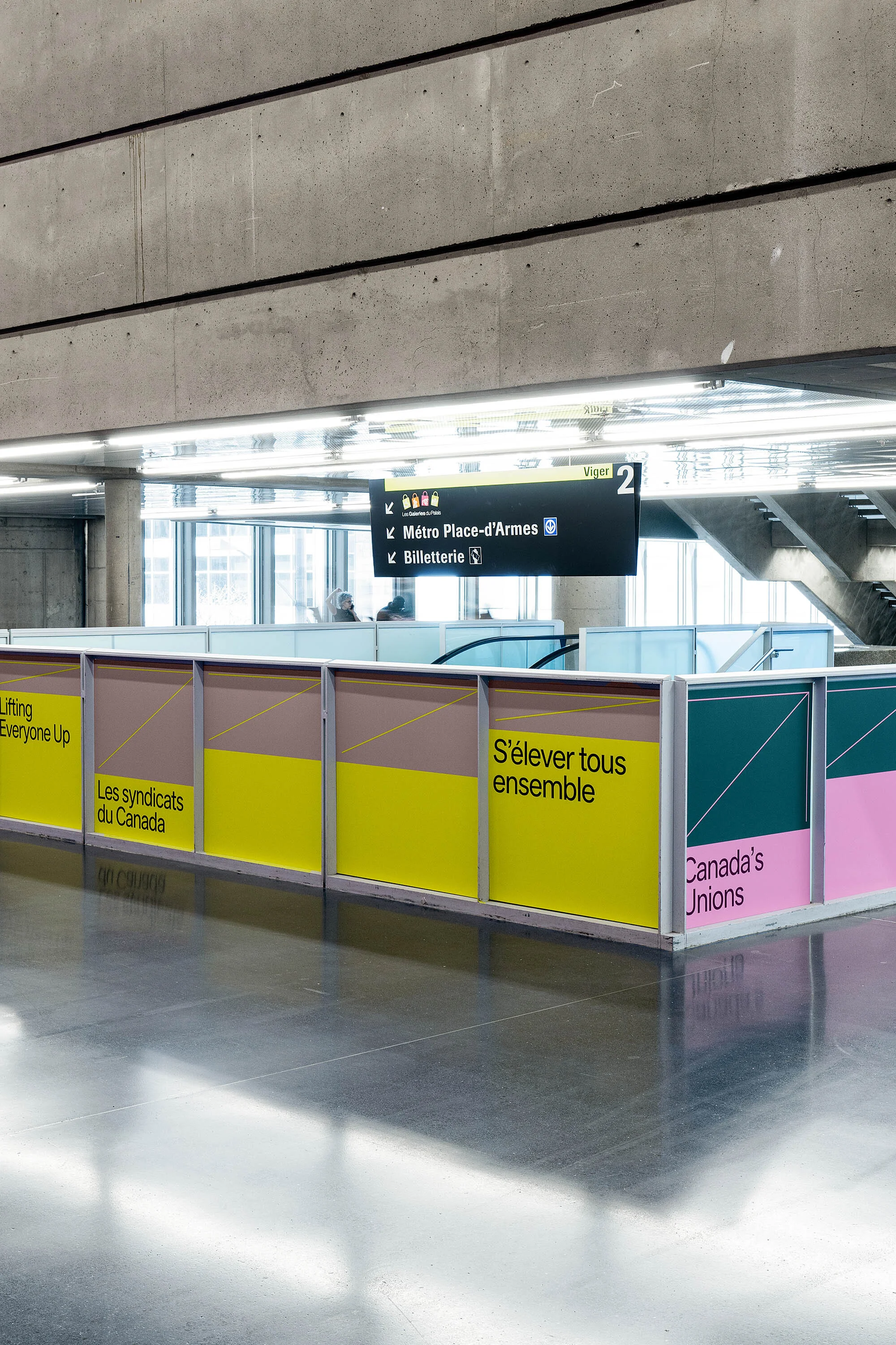 Indoor public transit station area with colorful graphic panels featuring text in both English and French, and overhead directional signs.