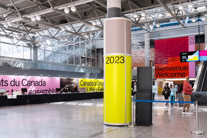 Spacious convention center lobby with event signage, structural column with '2023' text, and attendees walking towards escalators.