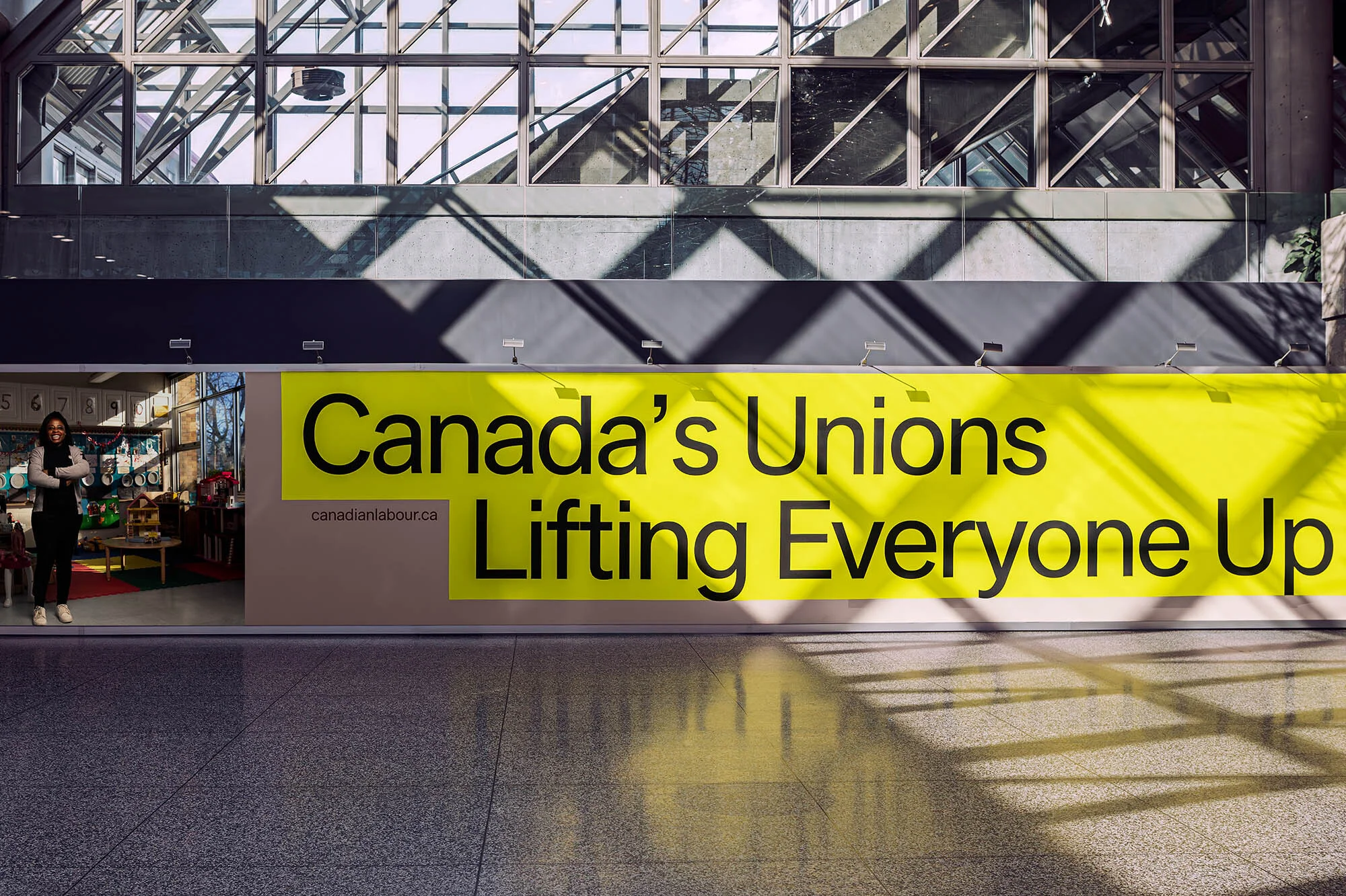 Indoor shot of a woman standing in front of a large yellow advertisement for Canada's Unions inside of a modern building.