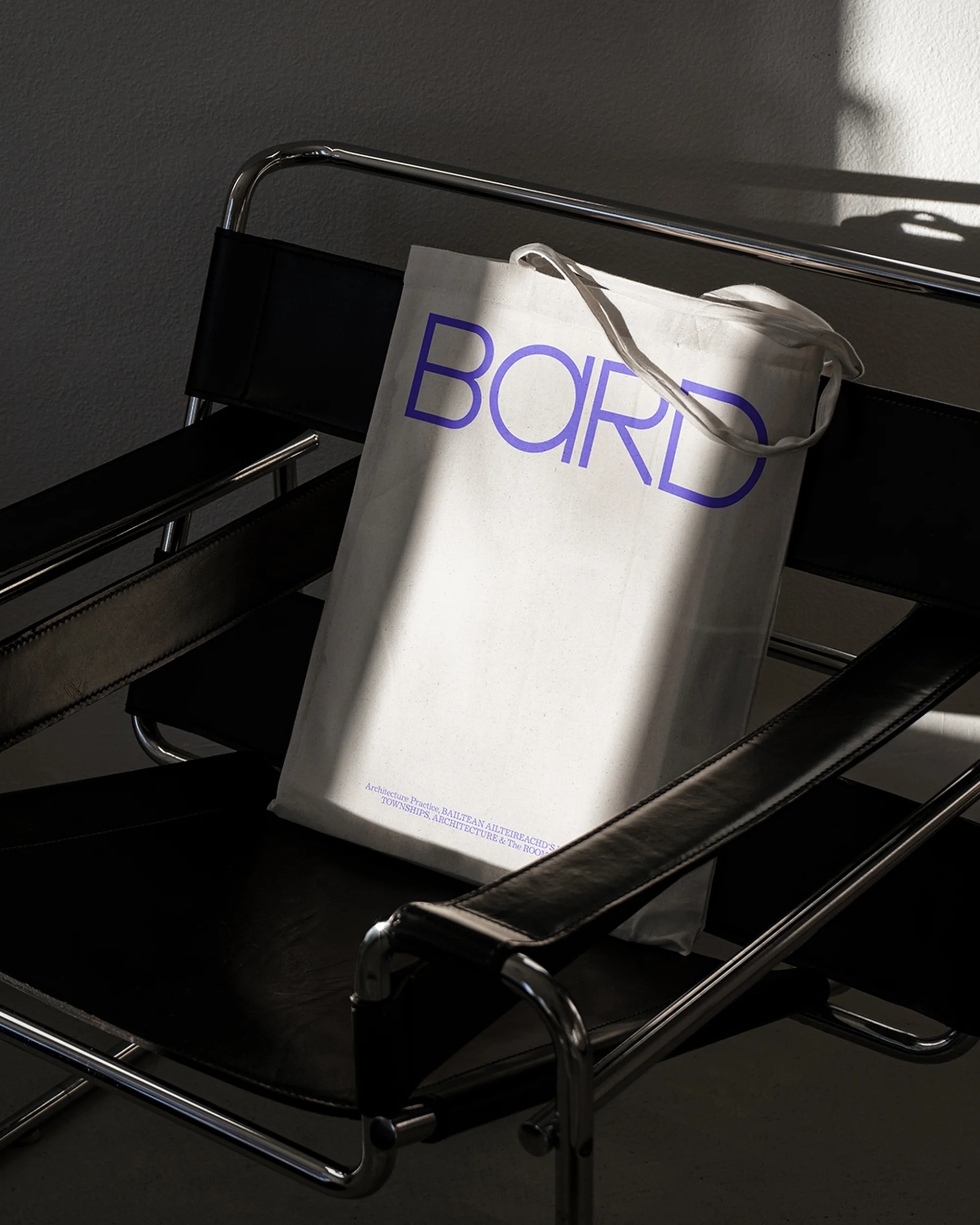 White tote bag with 'BARD' in blue, resting on a black chair, lit by natural light against a neutral wall.