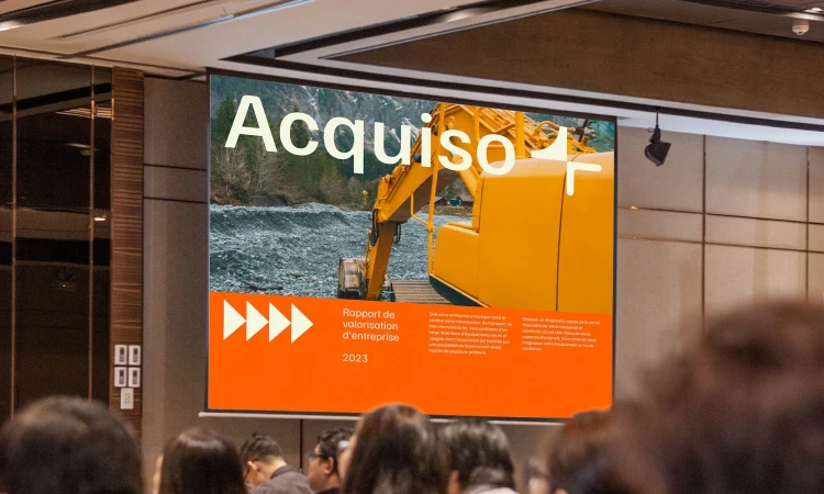 Presentation slide displaying a yellow excavator on a riverside, titled 'Acquiso', during a business meeting with attendees.