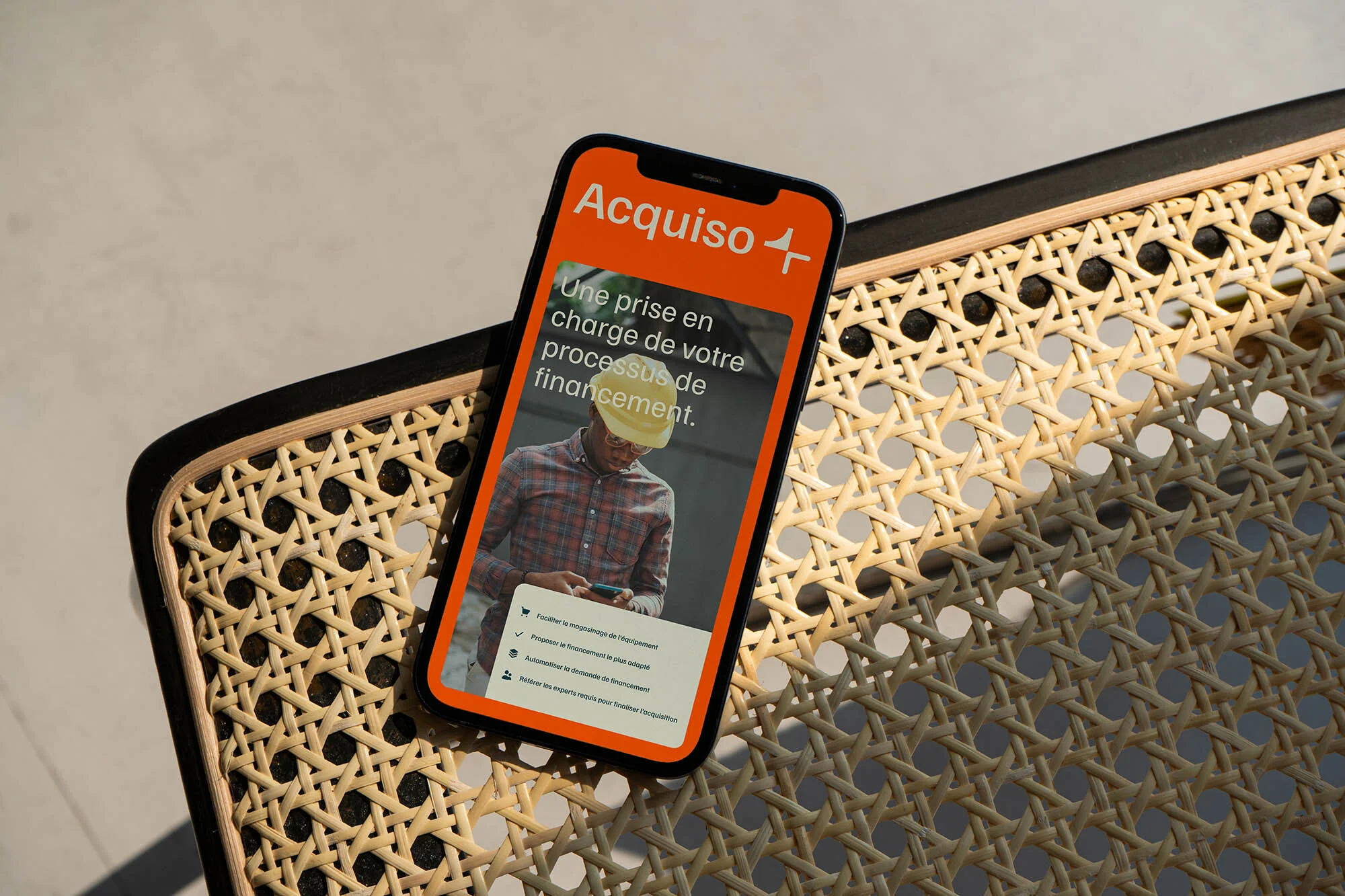 Orange mobile phone display showing Acquiso app, featuring a construction worker wearing a yellow hard hat on a wicker chair.