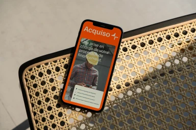 A smartphone resting on a woven rattan chair displaying the Acquiso brand mobile app interface with an orange background
