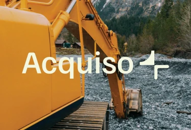 Yellow excavator arm against a rocky riverbed with pine trees and small wooden buildings in the background