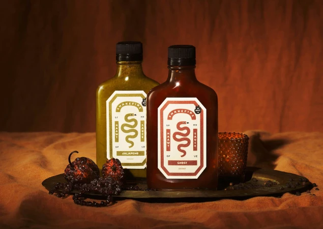 Two bottles of hot sauce, one green jalapeno and one red ghost pepper, sit with dried peppers on a dark metal tray against an orange backdrop.