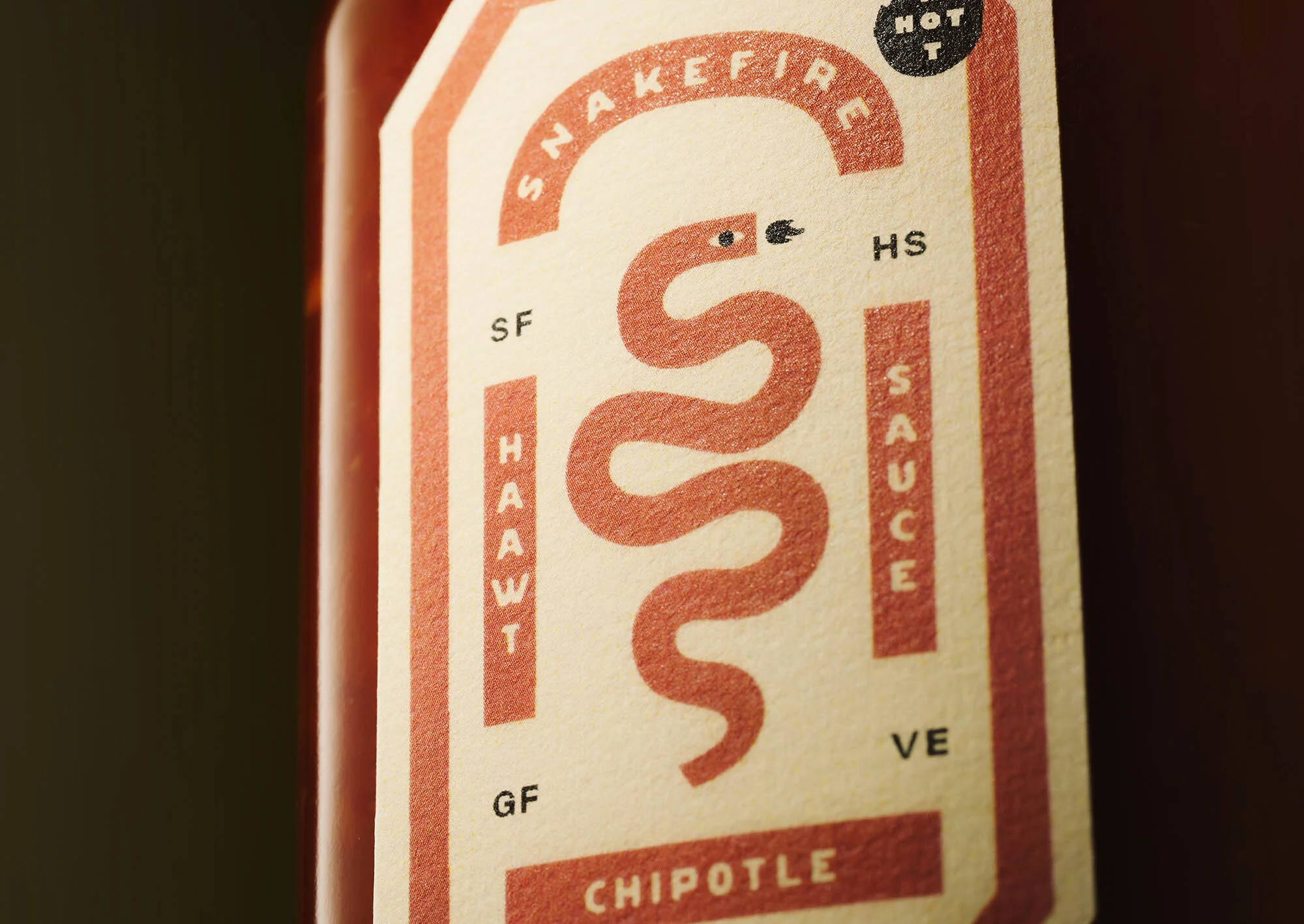 Close-up of 'Snakefire' chipotle hot sauce label with a stylized snake design and bold typography on a light beige background.