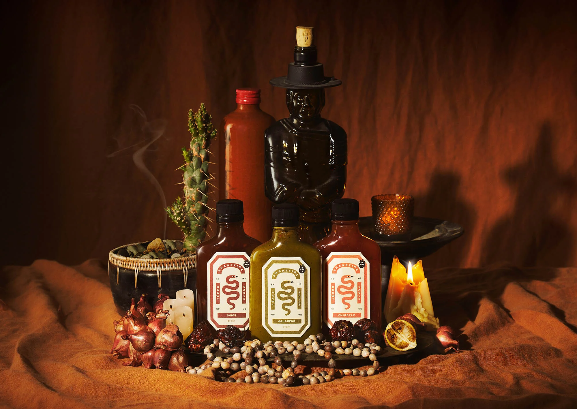 Several bottles of hot sauce and decorative elements are displayed, including a cactus, statue, candles, and dried fruit on a draped table.