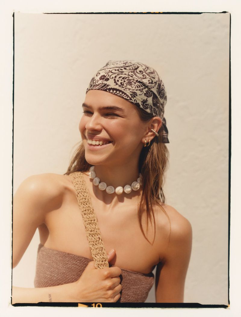 Smiling woman wearing a patterned headscarf, necklace, and brown strapless top against a white background in a medium shot.