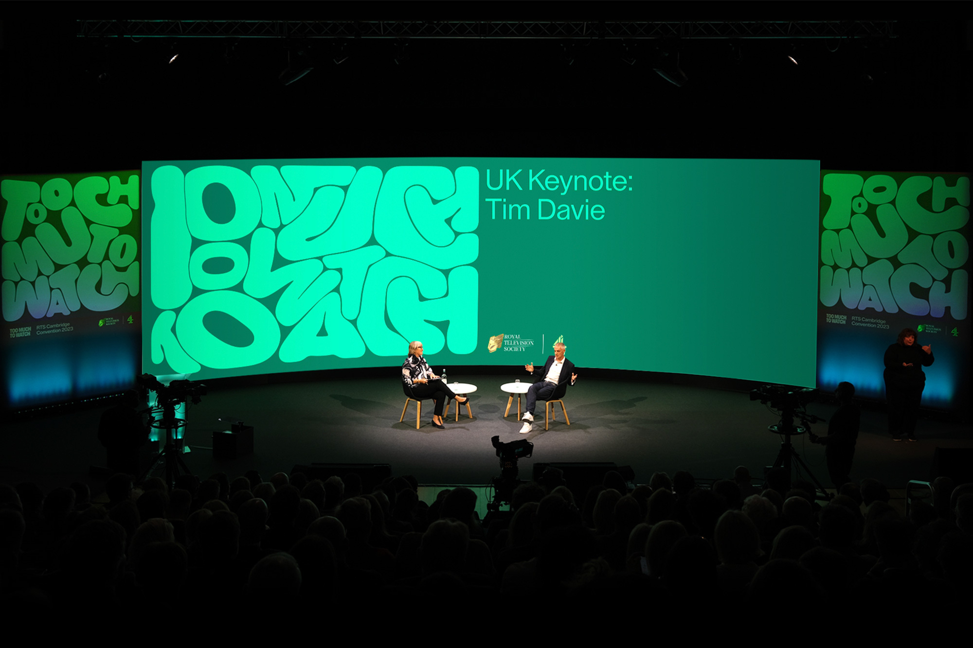 Two speakers sit on a stage before a large crowd in a dimly lit auditorium with a brightly lit screen in the background.