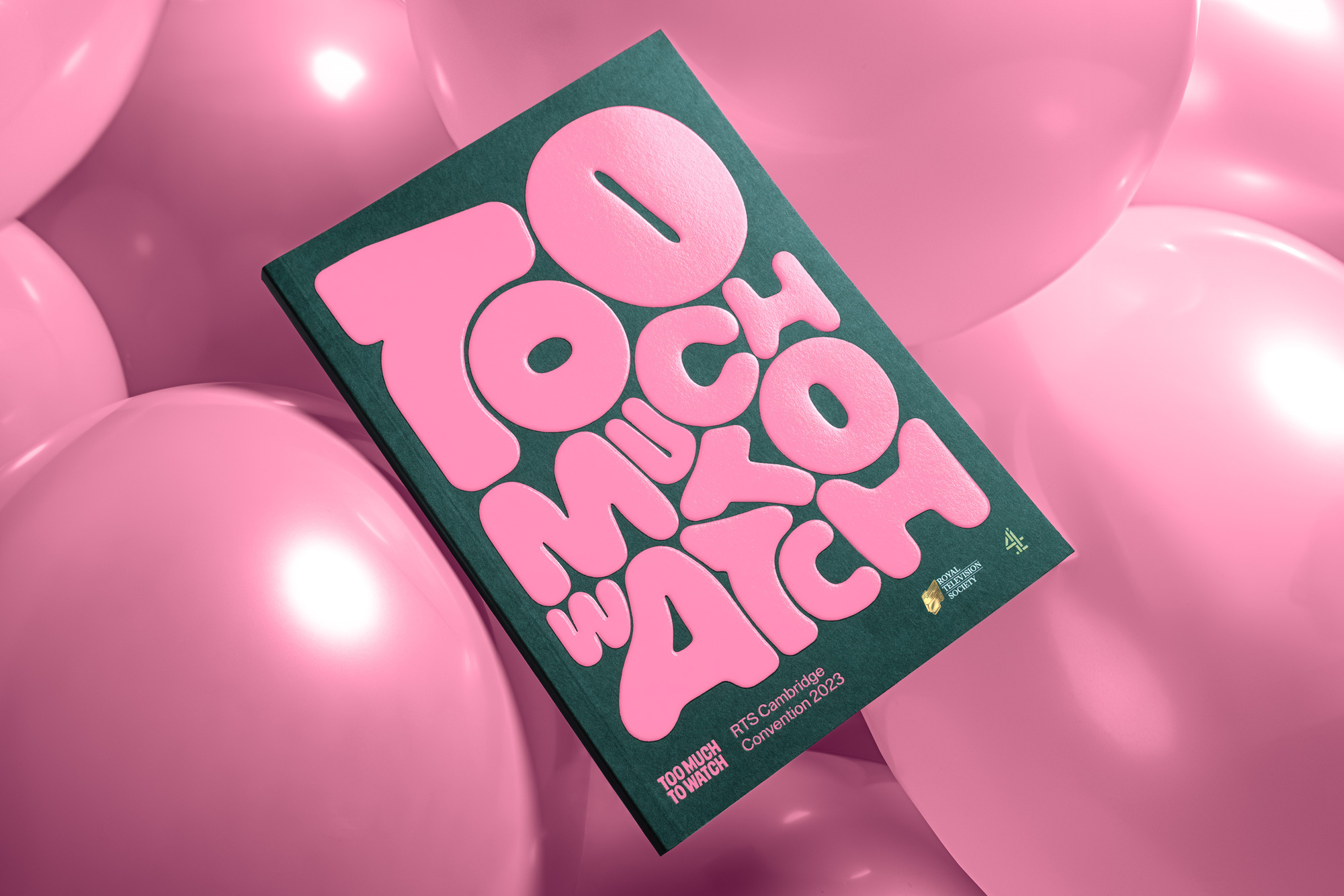 A studio shot shows a magazine cover with the pink text 'Too Much to Watch,' surrounded by multiple pink balloons, set against a green background.