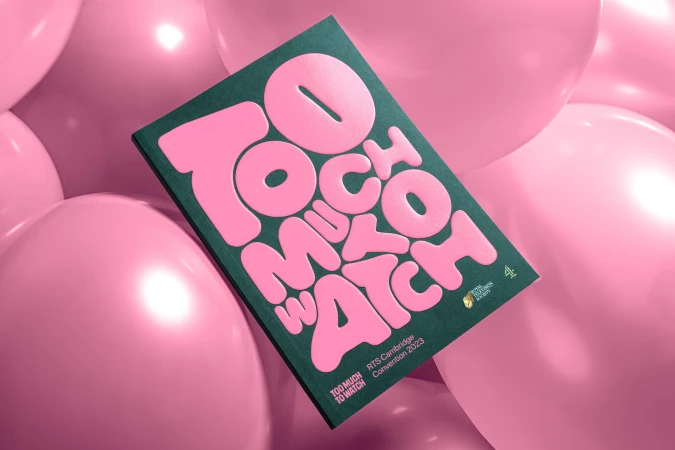 A studio shot shows a magazine cover with the pink text 'Too Much to Watch,' surrounded by multiple pink balloons, set against a green background.