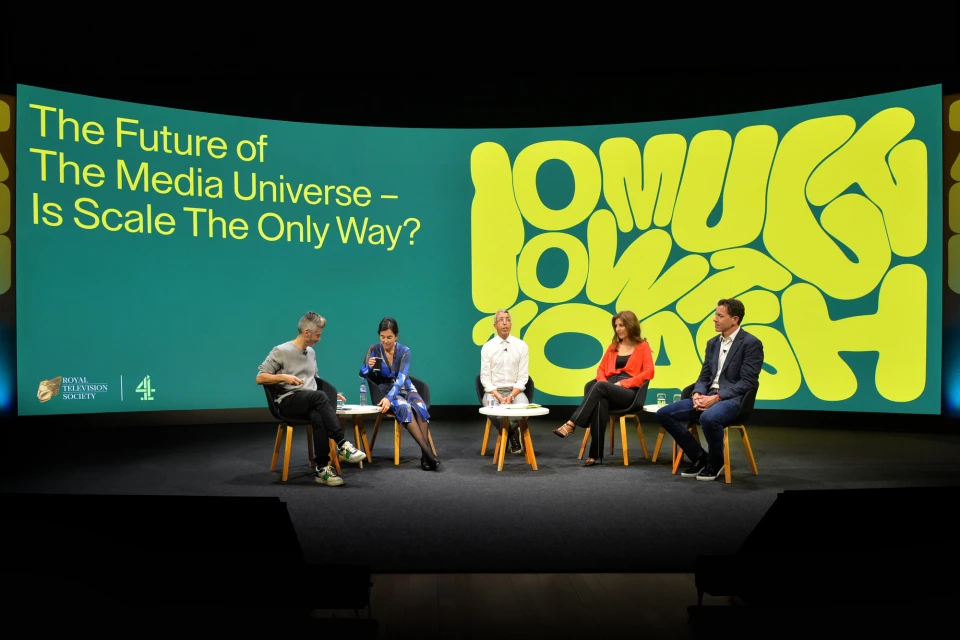 Panel discussion with five adults on stage in front of a green screen featuring graphic text about the media universe and scale.