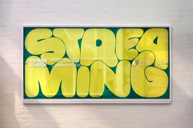 Indoor shot of a poster in a display case with the word 'STREAMING' written in yellow with green borders on a white brick wall.