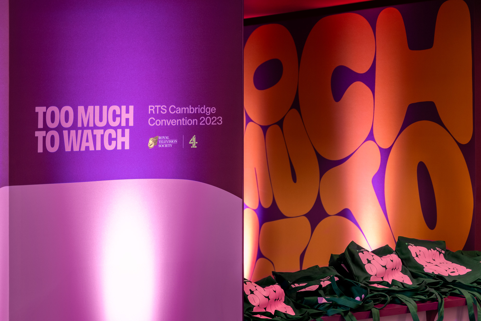 Indoor scene featuring a purple backdrop with white lettering for an event, with tote bags in the foreground.