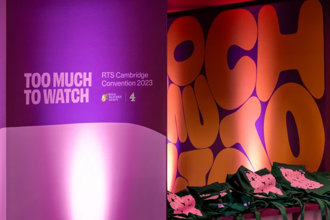 Indoor scene featuring a purple backdrop with white lettering for an event, with tote bags in the foreground.