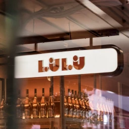 Illuminated storefront sign reading LULU above shelves displaying multiple wine bottles inside a commercial venue