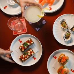 Overhead shot of a table with dishes including sushi, oysters, and toast, with hands holding a cocktail glass and a plate.