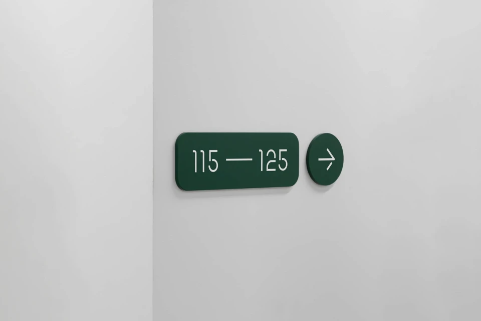 Dark green sign on a white wall indicating room numbers 115 to 125, with a right-pointing arrow indicating direction; simple modern design.