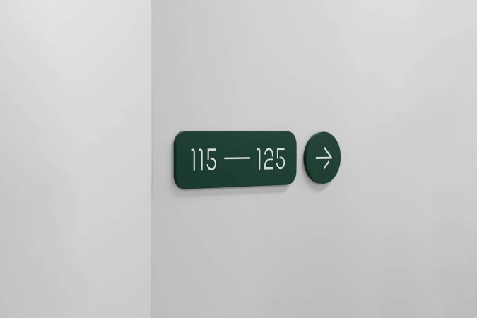 Dark green sign on a white wall indicating room numbers 115 to 125, with a right-pointing arrow indicating direction; simple modern design.