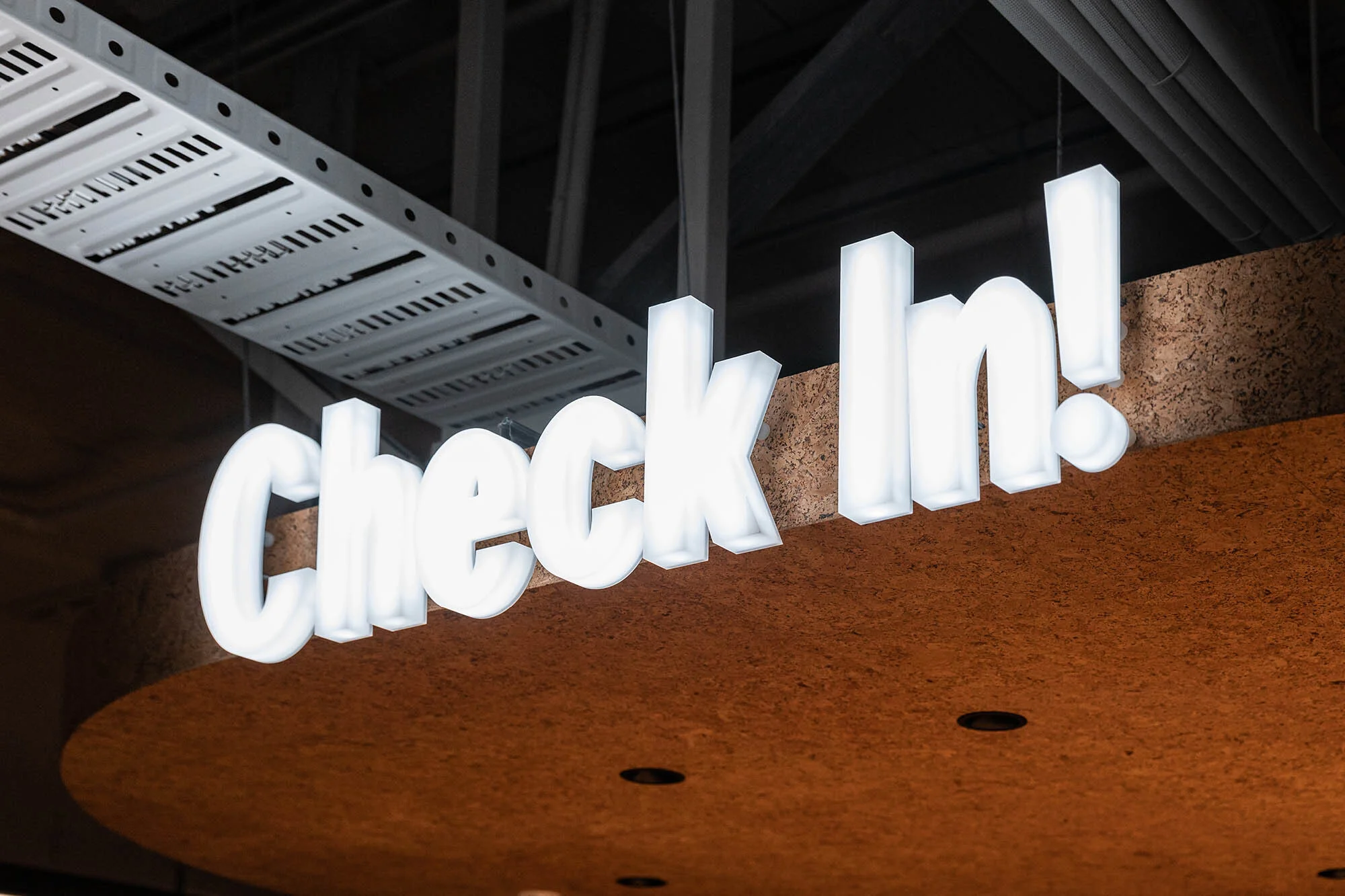 Illuminated 'Check In!' sign with white lettering against a brown textured surface and a gray ceiling with exposed piping.