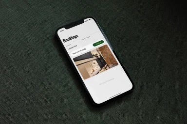 Black smartphone displaying a booking app interface resting on a dark green textured fabric surface.