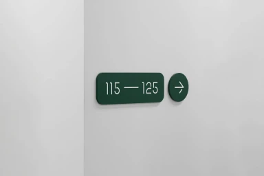 Dark green directional sign reading numbers 115 to 125 with an arrow icon mounted on a plain white wall