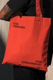 A person wearing black clothing carries a vibrant red canvas tote bag printed with the black text LESS ORDINARY. MORE EXTRAORDINARY.