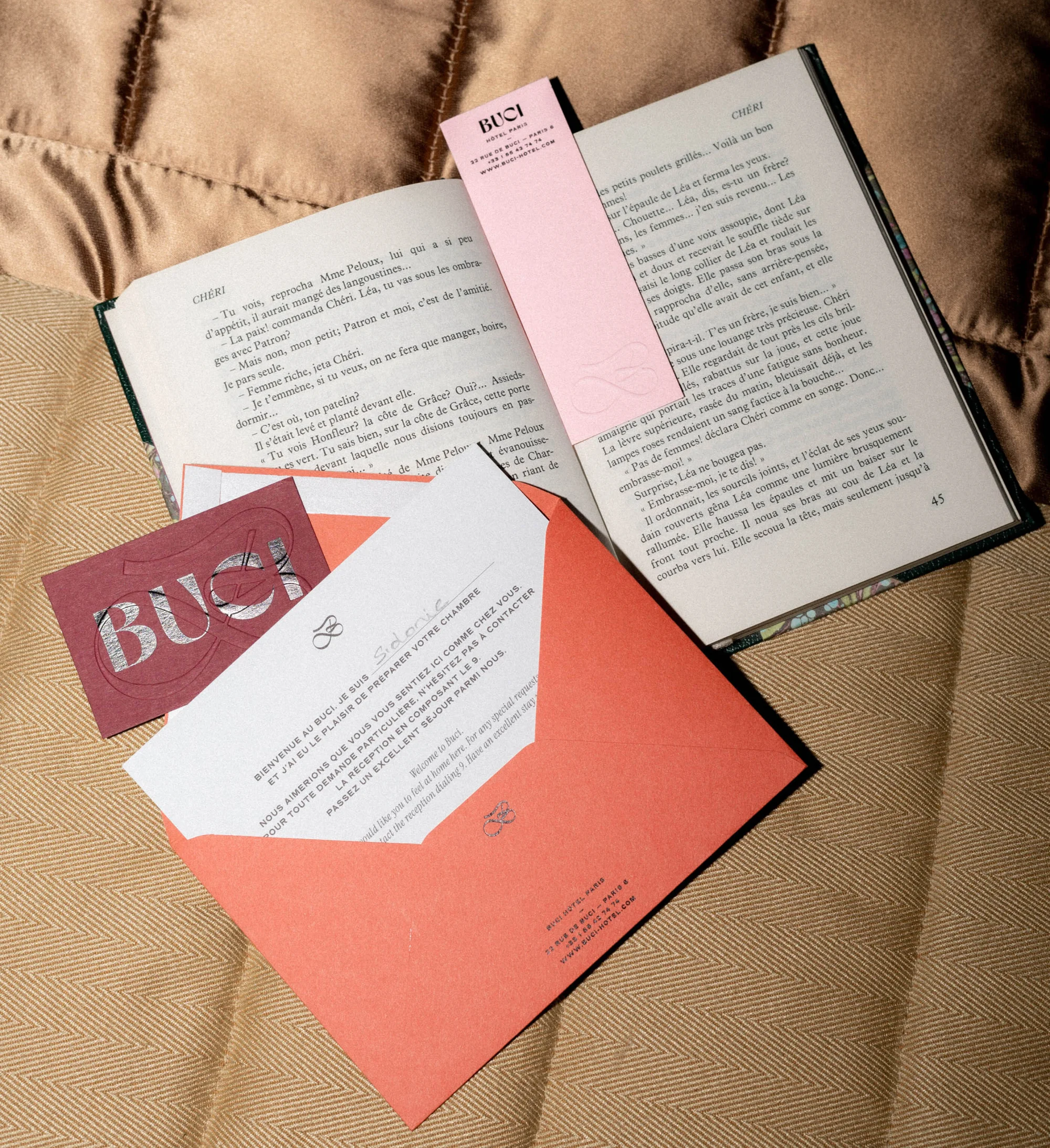 Open book with a pink bookmark, card, and salmon colored envelope on a tan quilted surface, inviting reader to enjoy.