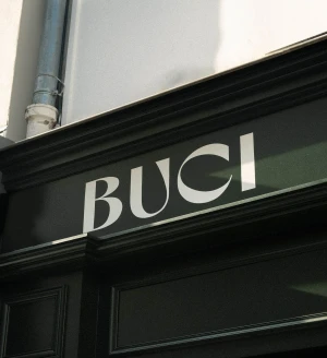 Signage featuring white lettering reading BUCI against a dark painted storefront facade under daylight.