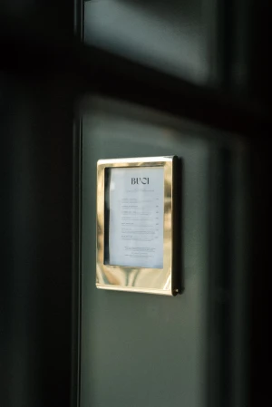 Close-up of a framed menu in a polished brass holder mounted on a dark green wall at Hotel Buci.