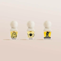 Three glass perfume bottles with rounded white caps and illustrated labels set against a neutral beige background